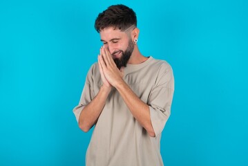 Overjoyed bearded caucasian man wearing casual T-shirt over blue background laughs joyfully and keeps palms pressed together hears something funny