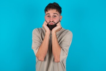 bearded caucasian man wearing casual T-shirt over blue background with surprised expression keeps hands under chin keeps lips folded makes funny grimace