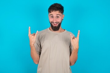 bearded caucasian man wearing casual T-shirt over blue background makes rock n roll sign looks self confident and cheerful enjoys cool music at party. Body language concept.