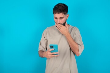 bearded caucasian man wearing casual T-shirt over blue background being deeply surprised, stares at...