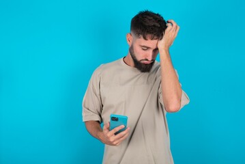 Upset depressed bearded caucasian man wearing casual T-shirt over blue background makes face palm as forgot about something important holds mobile phone expresses sorrow and regret blames