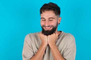 Fototapeta premium Cheerful bearded caucasian man wearing casual T-shirt over blue background has shy satisfied expression, smiles broadly, shows white teeth, People emotions