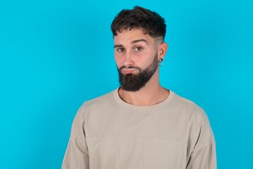 Obraz premium Displeased bearded caucasian man wearing casual T-shirt over blue background frowns face feels unhappy has some problems. Negative emotions and feelings concept