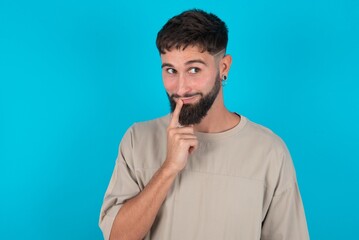 Lovely dreamy bearded caucasian man wearing casual T-shirt over blue background keeps finger near lips looks aside copy space.