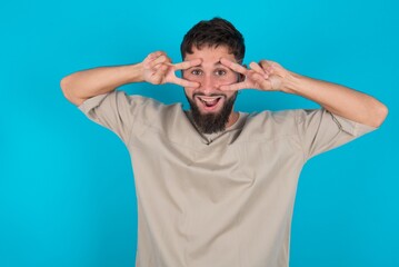 Cheerful positive bearded caucasian man wearing casual T-shirt over blue background shows v-sign near eyes open mouth