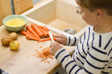 A happy Caucasian boy of 9 years old cleans a carrot in the kitchen with a safety knife. Assistance to parents, development through everyday activities.