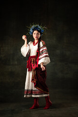 Adorable woman wearing national folk Ukrainian attire posing isolated over dark vintage background. Fashion, beauty, cultural heritage, eras comparison