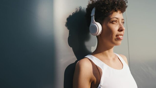 Pleased young African sporty woman listening music in headphones and looking at the camera outdoors