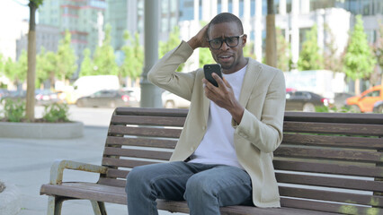 African Man Reacting to Loss on Smartphone while Sitting Outdoor on Bench