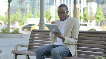 Fototapeta premium African Man Celebrating Online Win on Tablet while Sitting Outdoor on Bench