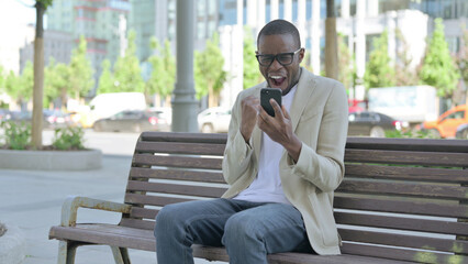 Fototapeta premium African Man Celebrating Online Success on Smartphone while Sitting Outdoor on Bench