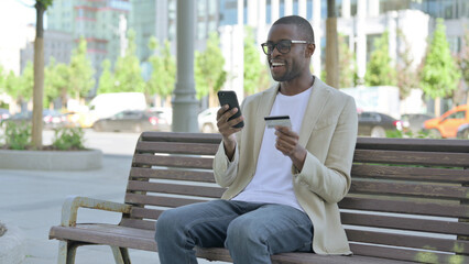 Fototapeta premium Excited African Man Shopping Online v on Phone while Sitting Outdoor on Bench