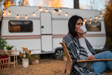 Caucasian woman in a knitted hat wrapped in a plaid and drinks a warming drink outdoors. Travel in a motor home in the fall.