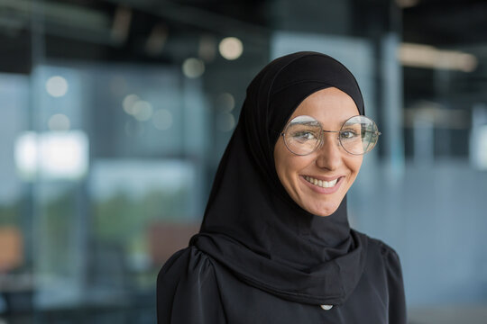 Close-up portrait of Muslim businesswoman in hijab smiling and looking at camera, female worker in glasses working inside modern office building.