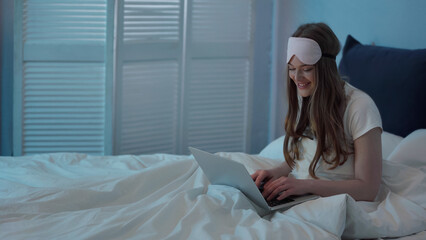 Cheerful woman in sleep mask using laptop on bed at night.