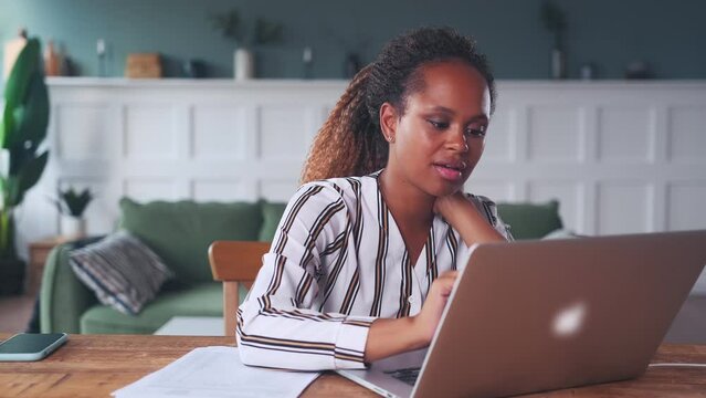 Young focused African American woman works with laptop and documents collating data and checking reports of employees with business indicators sits at work-table in own apartment. Manager, secretary