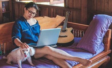 Young modern Asian woman in a casual outfit and eyeglasses sitting at a table with a notebook and working on a laptop while a cute small dog is beside her. work from home