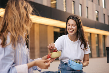 Two young nice caucasian girls enjoy eating sitting outside near city building in summer. Brunette...