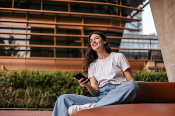 Pretty young caucasian woman holding smartphone, relaxing sitting outdoors. Brunette wears white t-shirt with jeans. Relaxed lifestyle, concept