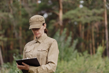 A woman biologist in uniform with a clipboard explore the forest biocenosis in national park in summer, selective focus. - obrazy, fototapety, plakaty
