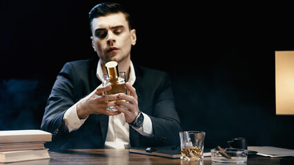 Businessman holding bottle of whiskey near books and cigar on table on black.