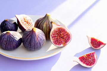 Whole and sliced purple figs on white ceramic plate. Fresh ripe fruits in the sunlight on lilac background. Close-up. Selective focus. Blurred background.