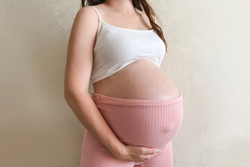 A pregnant woman in pink shorts and a white top holds her hands on her stomach on a white background. The concept of pregnancy, motherhood, preparation and expectation. Close-up, copy space.