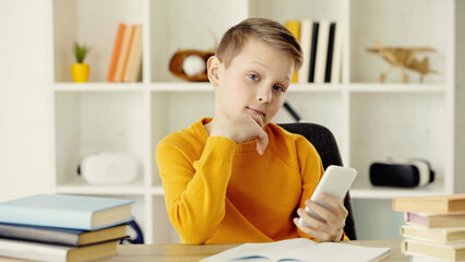 pensive schoolboy holding mobile phone near copy book and books on desk.