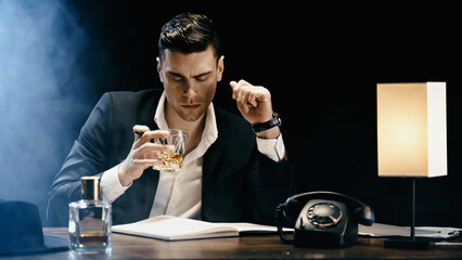 Businessman holding whiskey near notebook and telephone on black background with smoke.