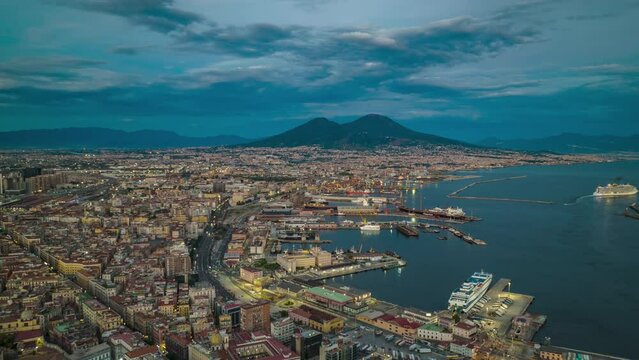 Twilight in coastal city. Forwards fly above famous city with port at sunset. Evening aerial panoramic hyperlapse shot of Naples and Mount Vesuvius massif in distance, Italy