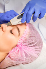 Fototapeta premium Young caucasian woman receiving facial skin cleaning by ultrasonic cosmetology face equipment in a medical salon