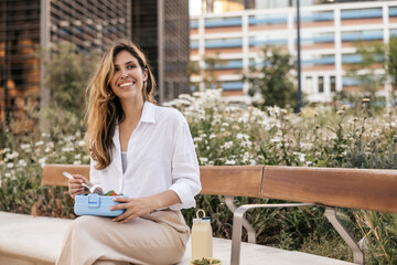 Smiling young caucasian business woman is sitting on bench outside and eating salad. Blonde smiles with her teeth, wears shirt. Free time concept
