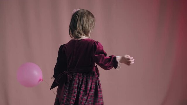Cute little girl in an vintage burgundy dress is playing with a pink inflatable balloon. The concept of children theater productions and developing acting circles. Fuchsia color background