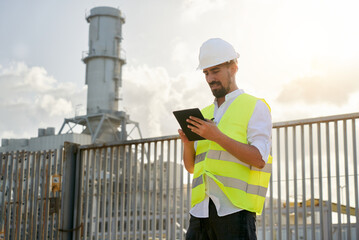 Photo of a young caucasian male engineer using digital table to do inspections at energy generator plant. Concept of a man outside workplace standing wearing protective equipment.