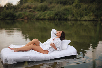 Brunette woman with short hair in a white shirt in bed on the water. A young woman floats on an inflatable mattress with a white sheet on the lake.