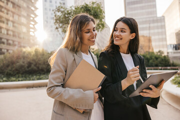 Cheerful caucasian young female colleagues with tablet communicate on work outdoors. Blonde and...