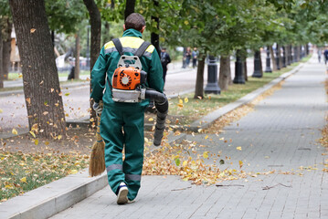 Man worker in green uniform clean the street with leaf blower. Falling leaves removal in autumn city