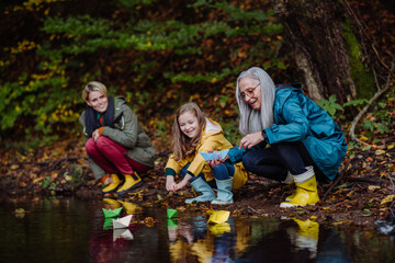 Small girl with mother and grandmother playing with paper boats in lake outoors in nature.