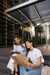 Two caucasian young female students are consulting about studying together sitting outdoors. Blonde...