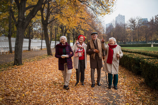 Group of happy senior friends with books on walk outdoors in park in autumn, talking and laughing
