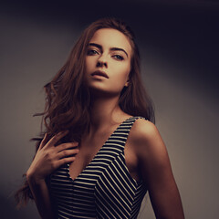 Fototapeta premium Beautiful romantic woman with nude natural makeup and long curly hair style posing in striped dress on grey background. Closeup portrait
