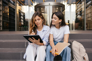 Calm young caucasian girls use tablet to search for information sitting on street. Blonde and...