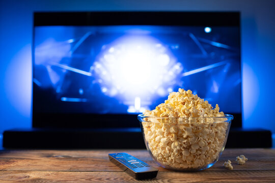 A glass bowl of popcorn and remote control in the background the TV works. Evening cozy watching a movie or TV series at home