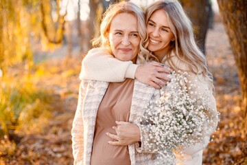 Adult daughter hugs her pregnant mom while standing in autumn park - obrazy, fototapety, plakaty