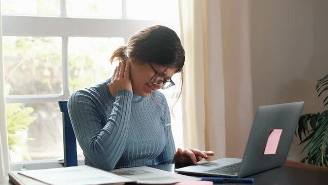 One young woman feeling bad and having pain for overstudying at home on the table. Neck ache concept. Portrait and close up of hands touching neck.
