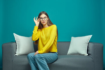 Pretty young girl student fixing her glasses, looking at camera, sitting on sofa. Advertising of...