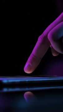 Vertical video. Young woman browsing internet on smart phone at night; girls finger tapping keypad, scrolling touchscreen of mobile device, purple blue low light; extreme close up side view