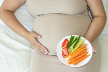 pregnant woman advanced belly third trimester lies on bed with plate with vegetables.cucumber carrots and tomatoes fruit.one hand caresses the belly fetus.awaiting mother,expecting child birth.healthy