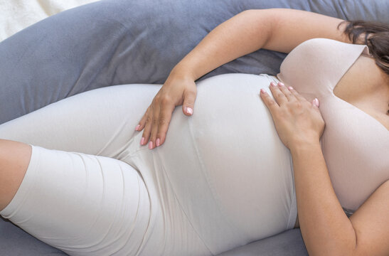 pregnant woman lies on bed in U shaped gray color pillow.happy future mother expecting baby awaiting period is resting relax in bedroom huggin the belly with both hands caressing with palms.third 