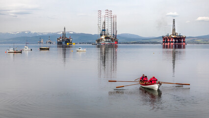 Rowers on sea at Cromarty on still water with sad and historical old oil rig graveyard along the...